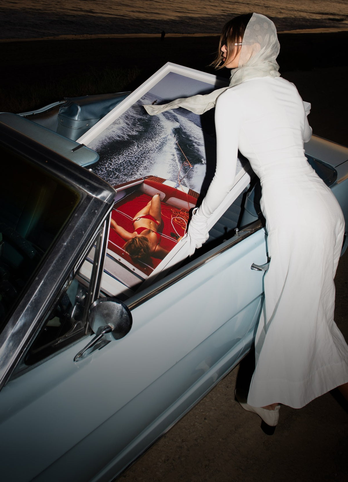 A woman in a white dress leaning over a vintage convertible at night, holding a framed Leisure Piece print